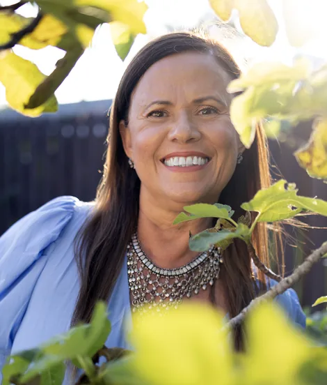 Isa Herrera outdoors in natural light, wearing a silver necklace, smiling directly at the camera with green foliage behind her.